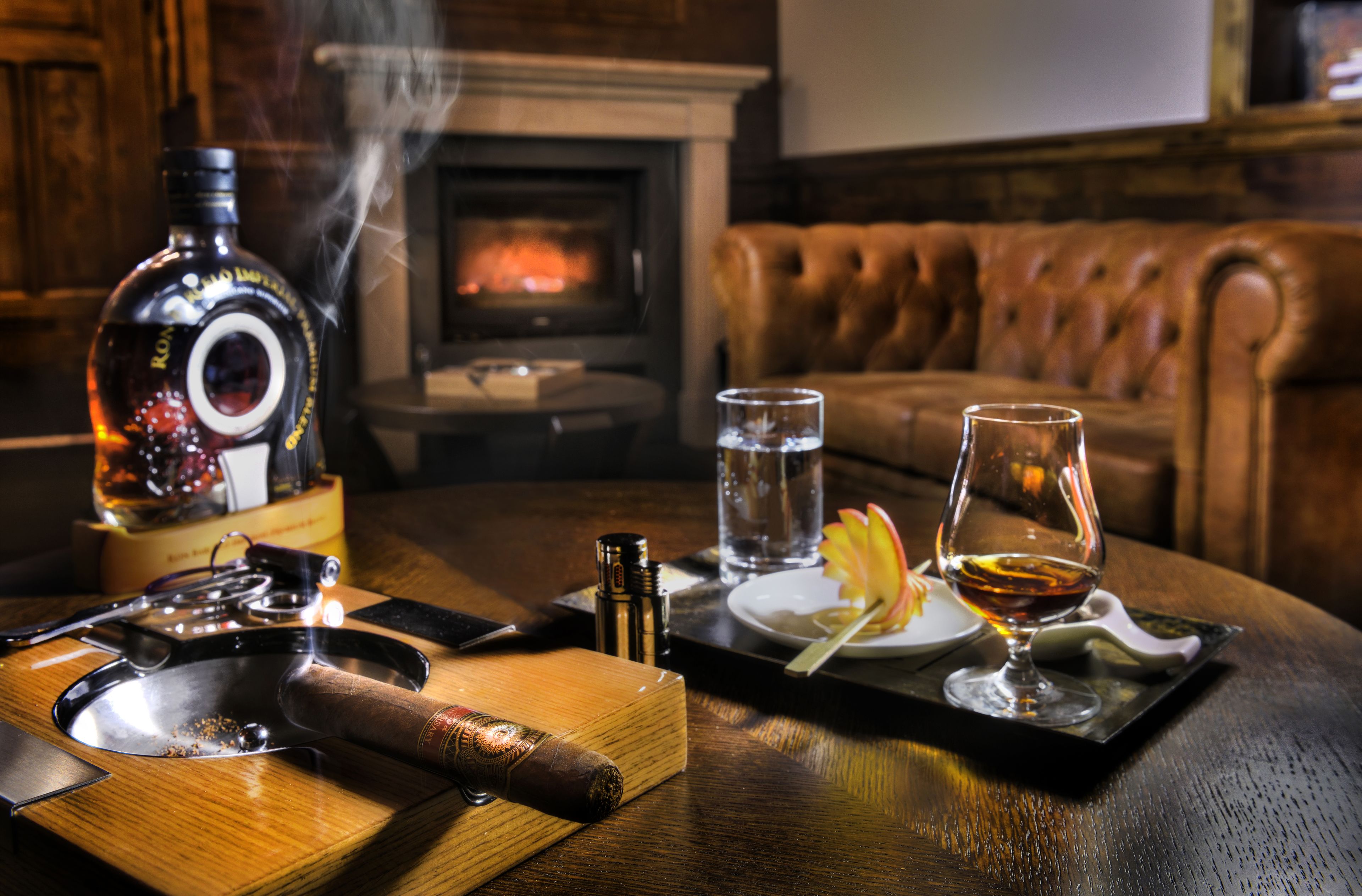 A wooden table with a cigar, a bottle of liquor, a glass of whiskey, and a tray with a glass of water and a cocktail glass.