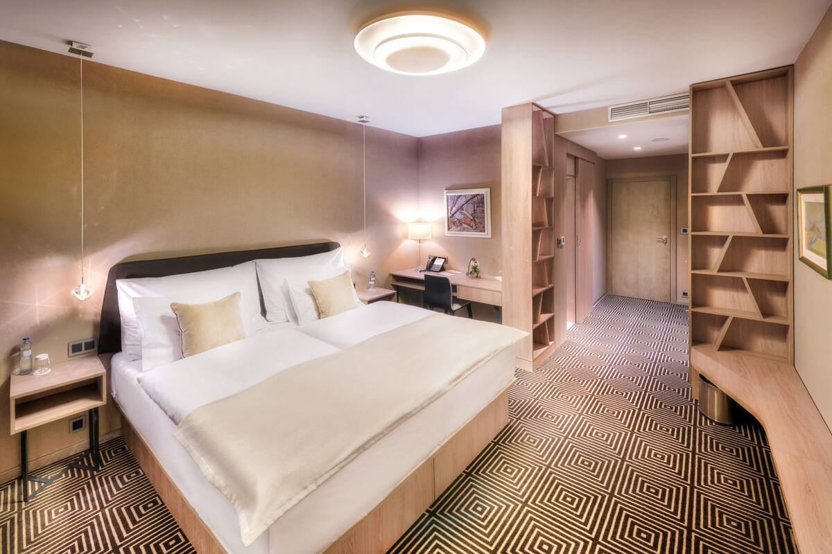 Een hotelkamer met een groot bed, nachtkastjes, een bureau, een kledingkast en een kamer met een patroon op de vloer.