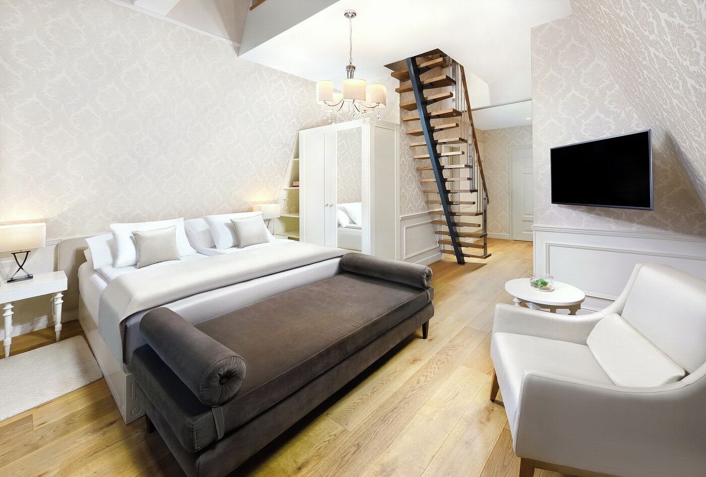 A modern bedroom with a large bed, a dark sofa, a white armchair, a TV mounted on the wall, and a wooden staircase.