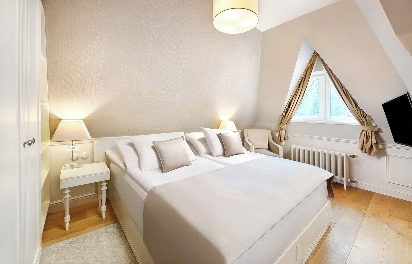 Een slaapkamer met een groot bed, nachtkastjes, een lamp, een radiator en een raam met gordijnen.