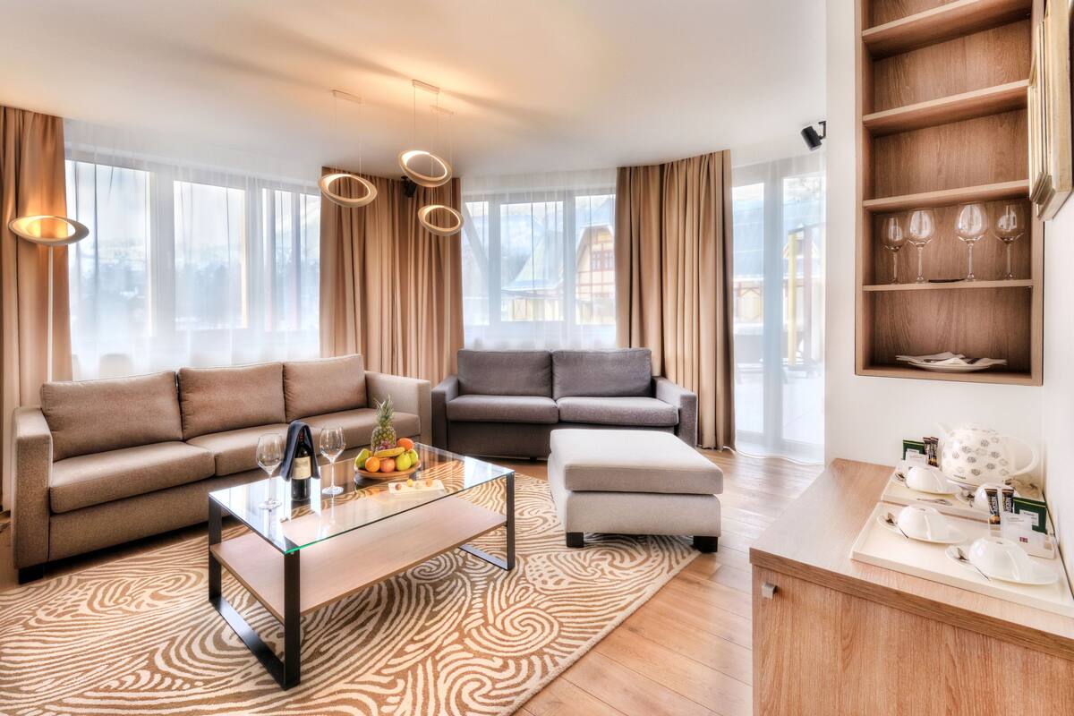 Een moderne woonkamer met een bank, voetenbankje, salontafel, een flatscreen televisie en een glazen tafel met fruit en wijn.