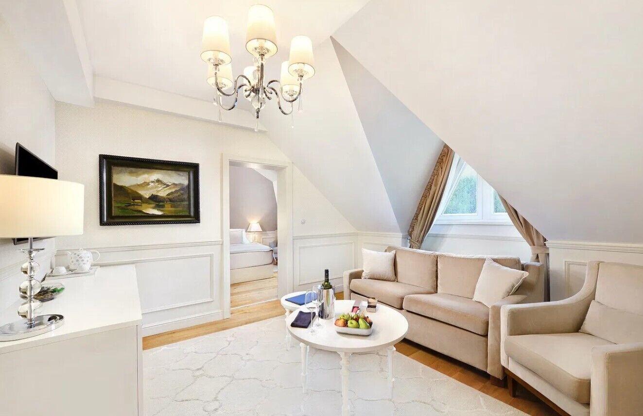 A modern living room with a high ceiling, a chandelier, a sofa set, a coffee table with fruits, and a painting on the wall.