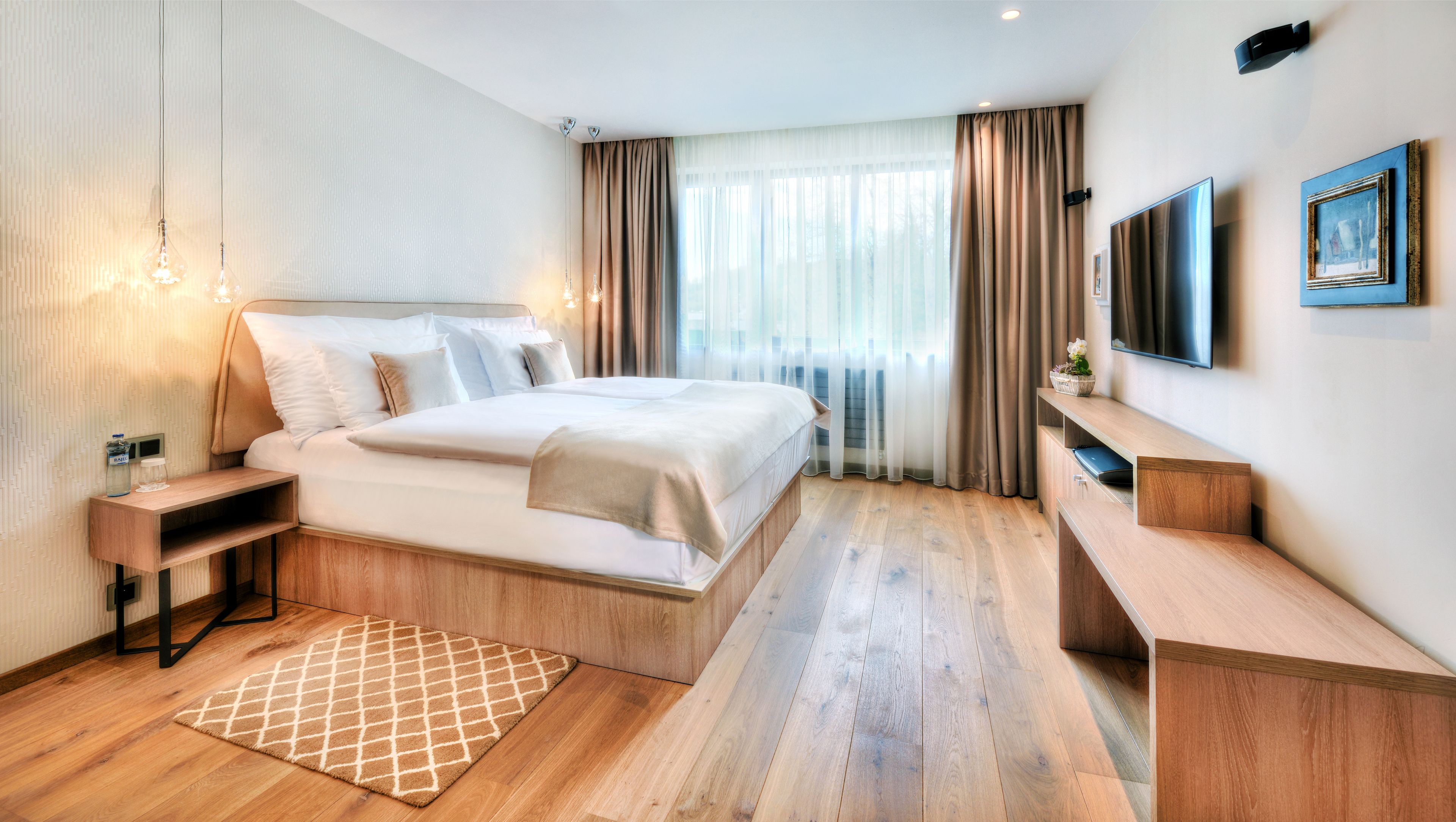 A modern hotel room with a large bed, bedside tables, a TV, and a wooden floor.