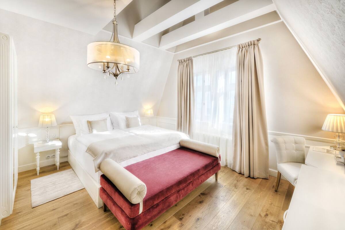 Een slaapkamer met een groot bed, een rode bedbank, een witte stoel, een nachtkastje en een kroonluchter.