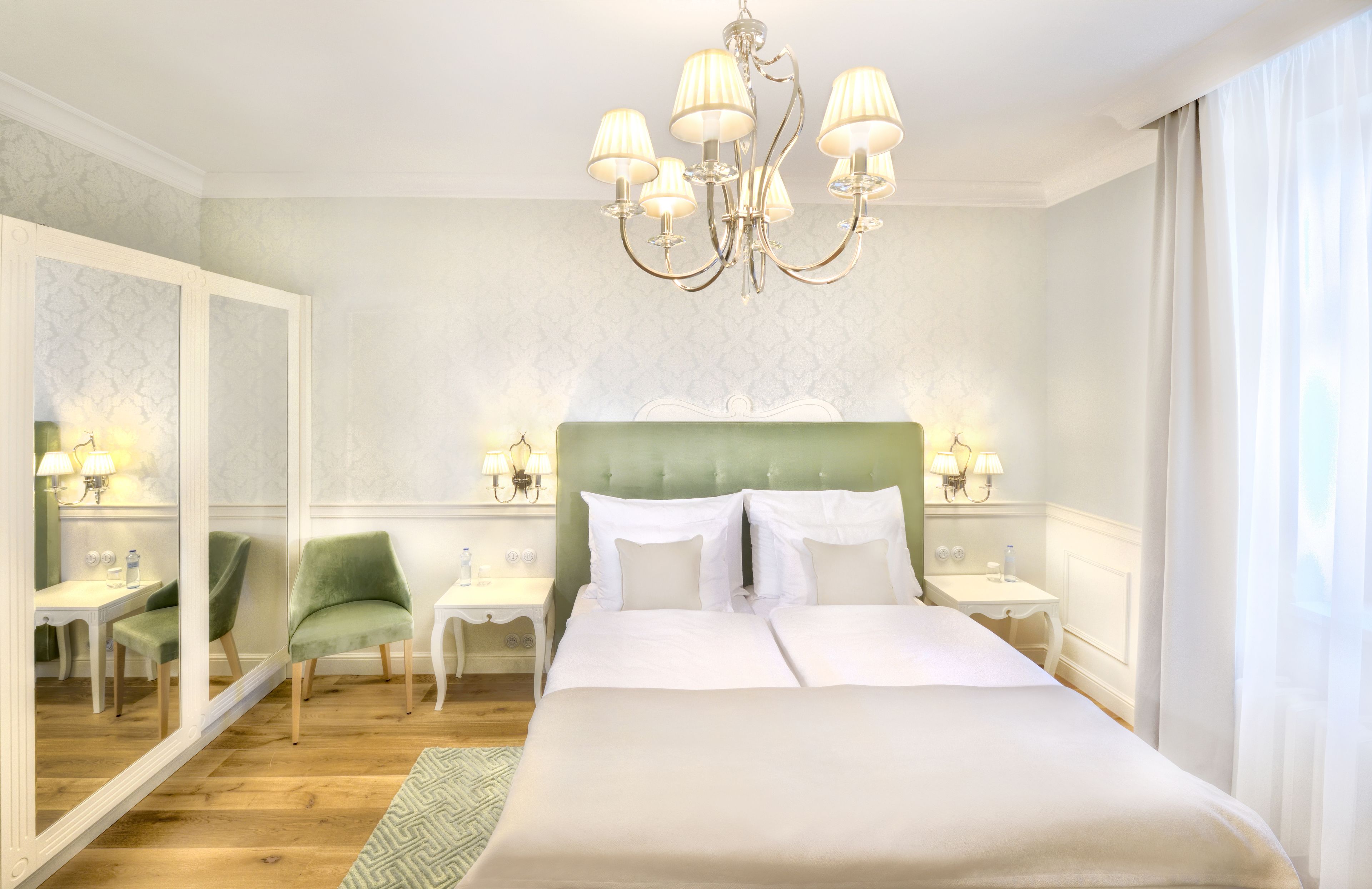A hotel room with a large bed, a green armchair, a small table, and a mirror.