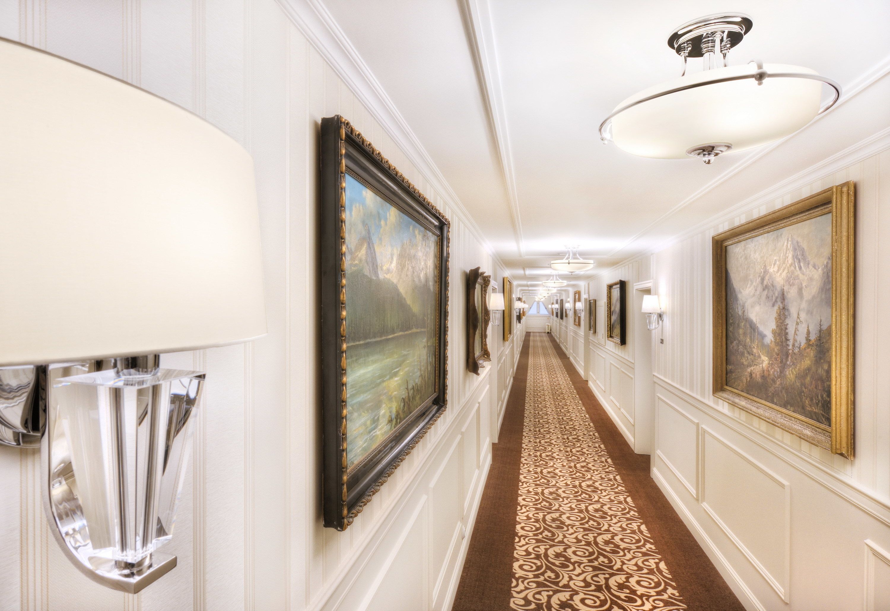 A hallway with framed artwork on the walls, a patterned carpet, and a chandelier.