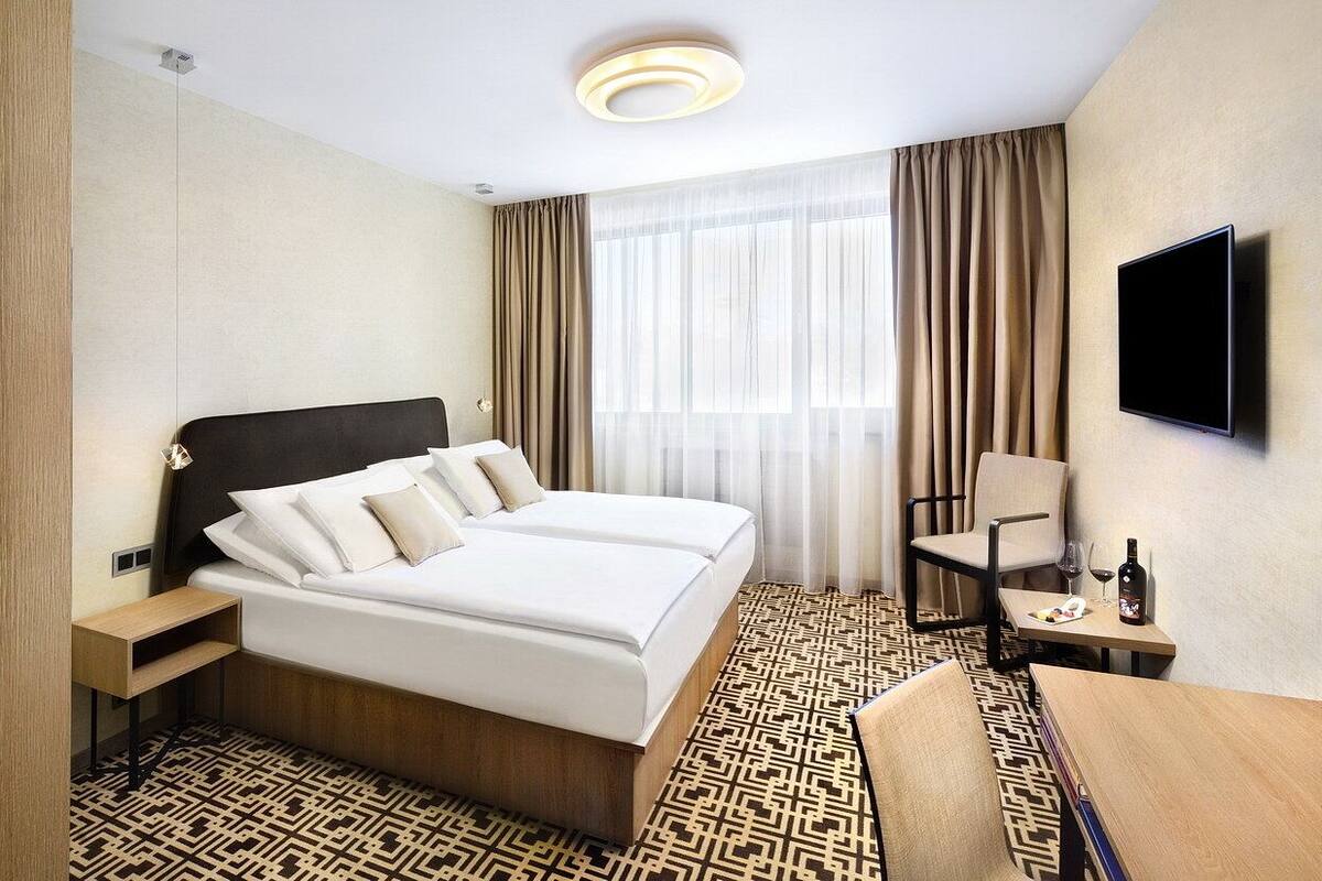 Een hotelkamer met een groot bed, een bureau met stoel, een aan de muur bevestigde televisie, en een raam met gordijnen.