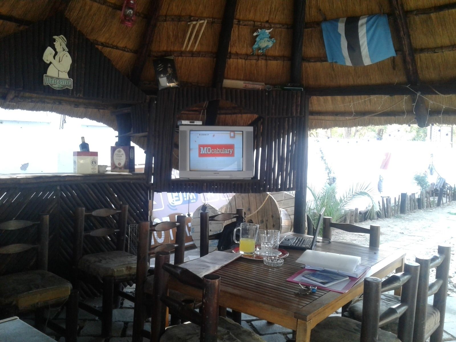 A rustic outdoor seating area with wooden tables and chairs, a television mounted on the wall, and a thatched roof structure.