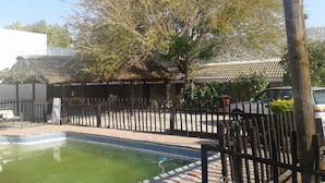 A swimming pool with a tiled edge, surrounded by a wooden fence, and a building with a tiled roof in the background. - Road View Lodge (Francistown)