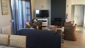 A living room with a sofa, two wicker chairs, a coffee table, a flat-screen TV, and a fireplace. - Pinnacle Point Pool Lodge (Mossel Bay)