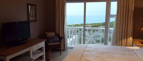 A bedroom with a bed, a TV, a desk, a chair, and a balcony with a view of the sea.