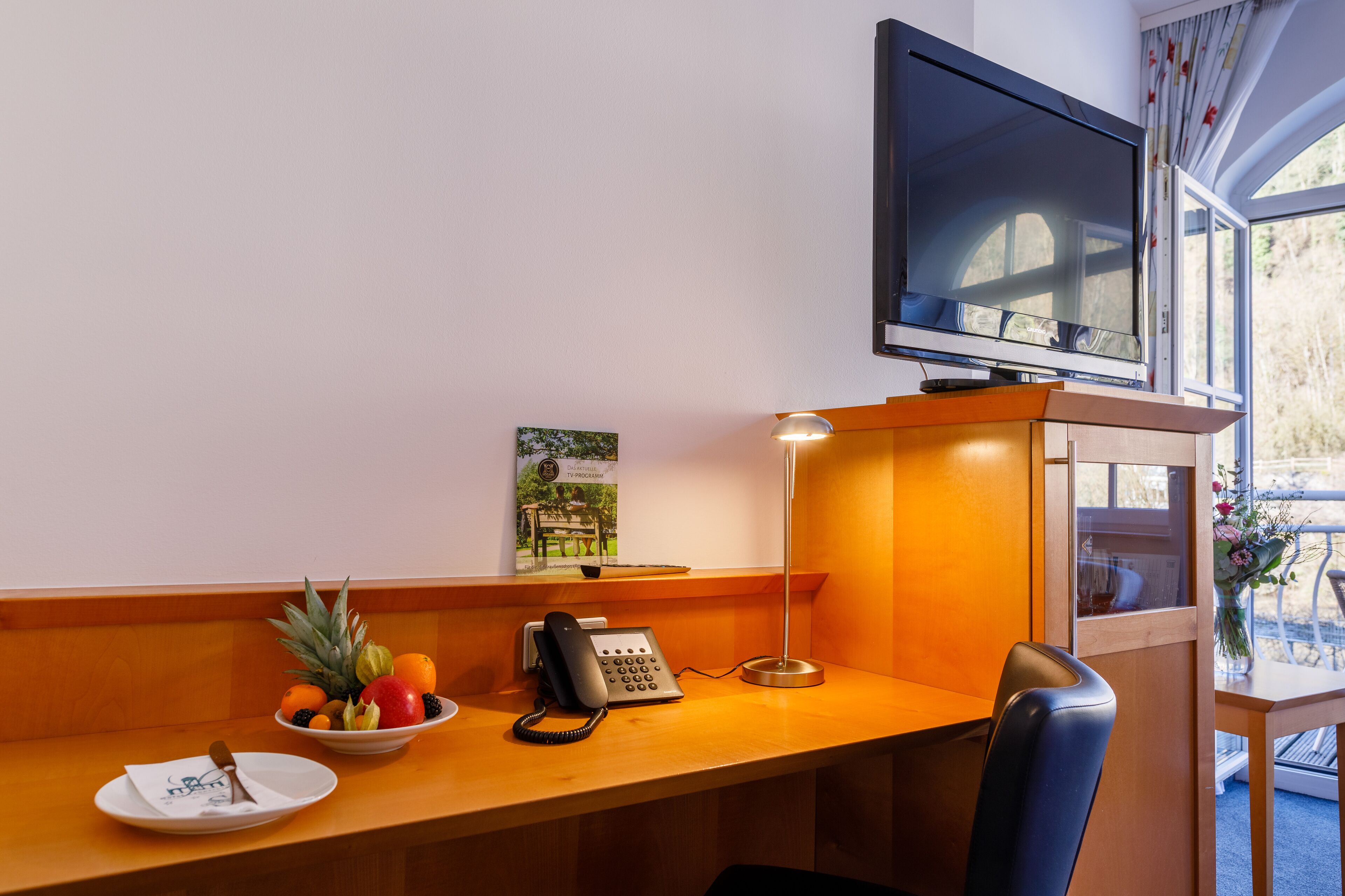 Habitación de hotel con un escritorio de madera, un teléfono, un frutero, un cuadro enmarcado y un televisor.