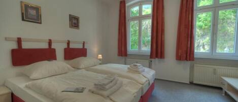 A hotel room with two beds, red headboards, white bedding, a nightstand, a window with curtains, and framed pictures on the wall.