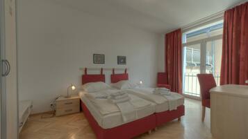 Cama doble con cabeceros rojos, una mesita de noche con lámpara, una silla y vistas al balcón exterior.