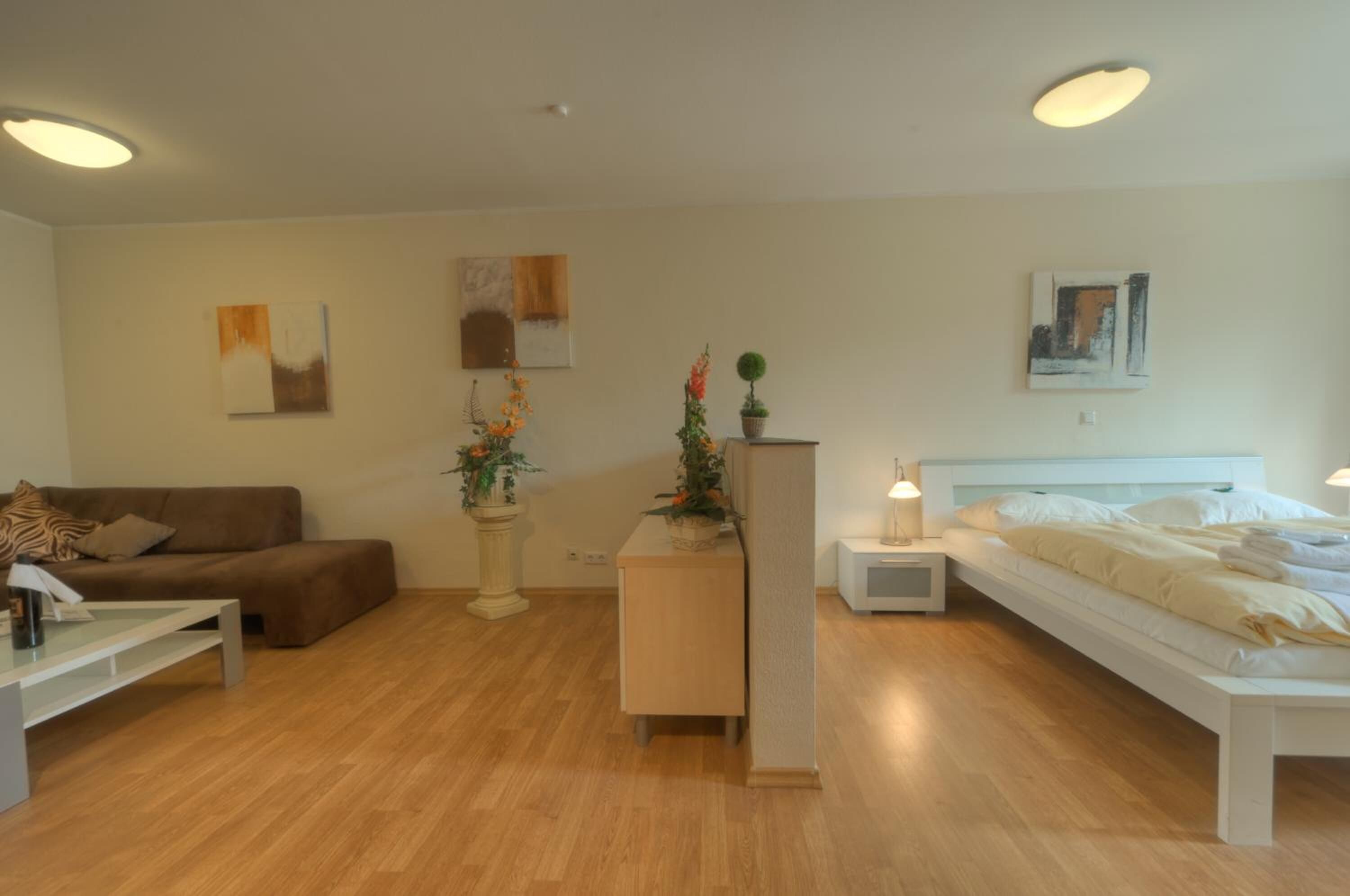 A modern, well-lit hotel room with a wooden floor, a white bed, a sofa, a small table, and two nightstands.