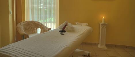 A spa room with a bed, a chair, a candle, and a painting on the wall.