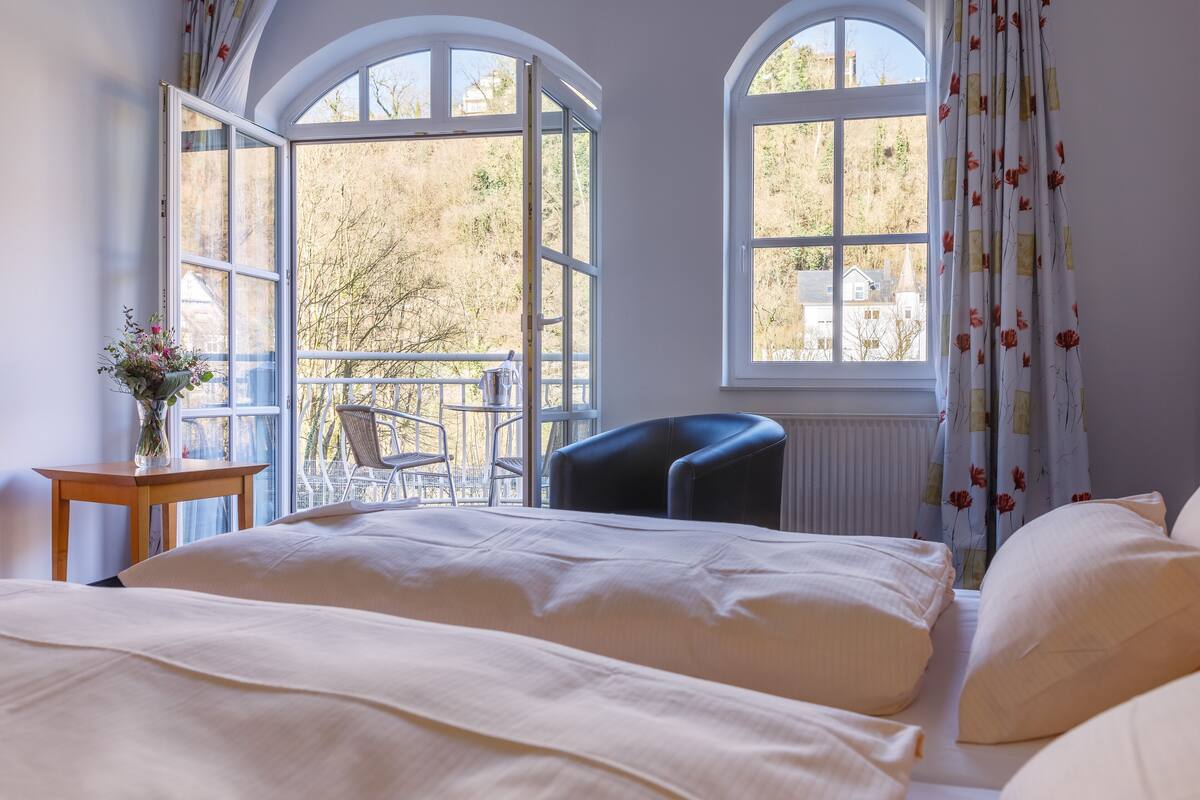 A bedroom with a bed, a chair, a table with a vase, and two windows.