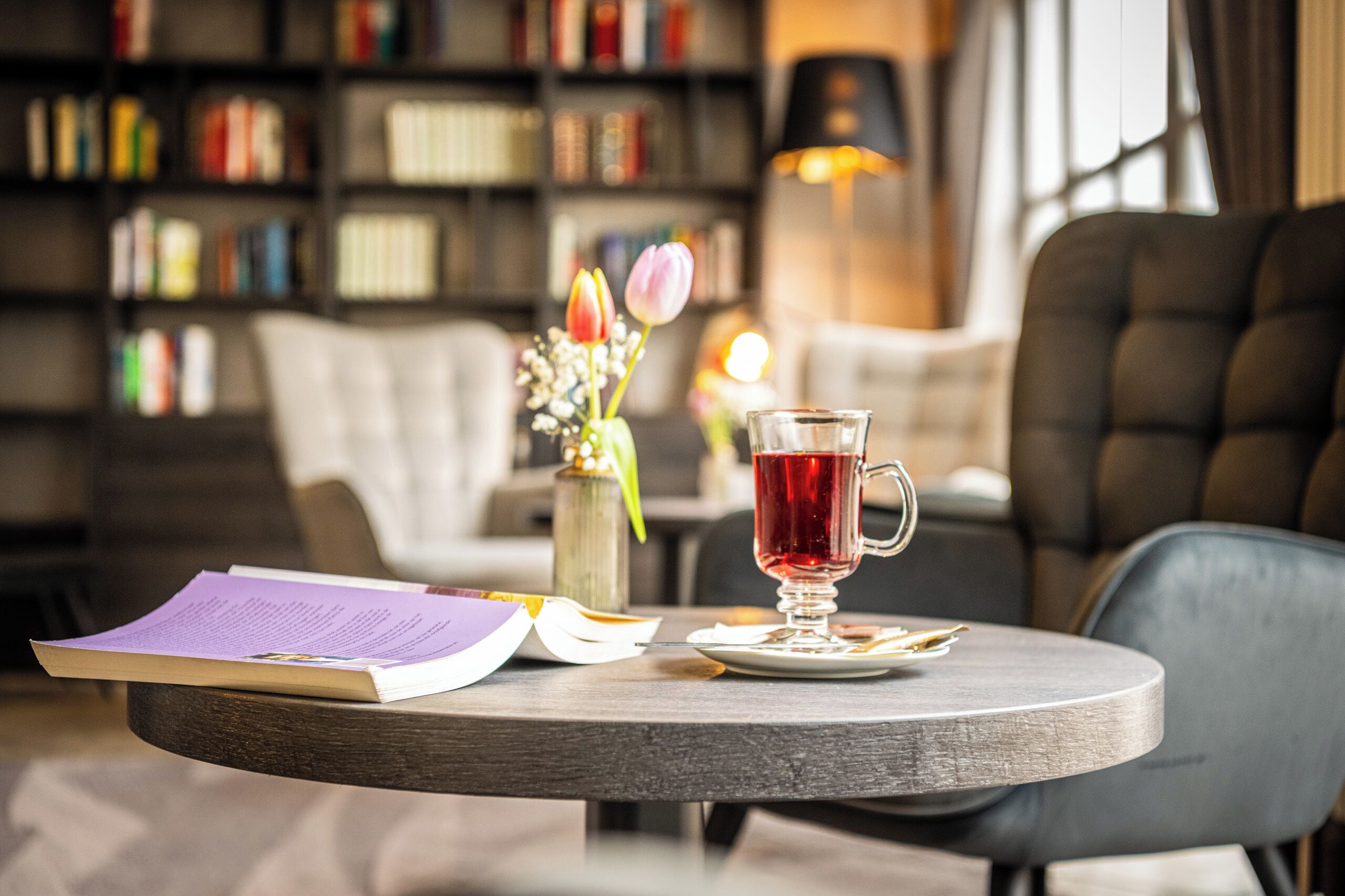 A round wooden table with a book, a glass of red beverage, and a vase with flowers.