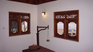 Una habitación con una mesa de masaje de madera, una balanza y dos ventanas con marcos de madera decorativos.
