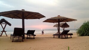 Una playa con sillones de madera y sombrillas de paja, un atardecer de fondo y una bandera a la derecha.
