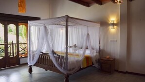 Una cama con dosel y marco de madera, con mosquitera blanca, una mesita de noche pequeña y una lámpara fijada a la pared.