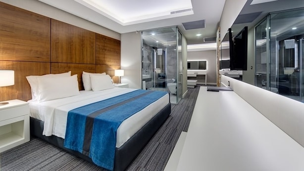Una habitación de hotel moderna con una cama grande, un escritorio, cabina de ducha con paredes de vidrio y un rincón con sofás.