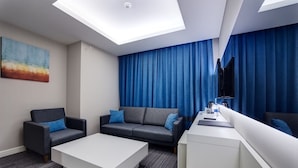 Una habitación de hotel moderna con sofá, una mesa de centro, un escritorio y un ventanal con cortinas azules.