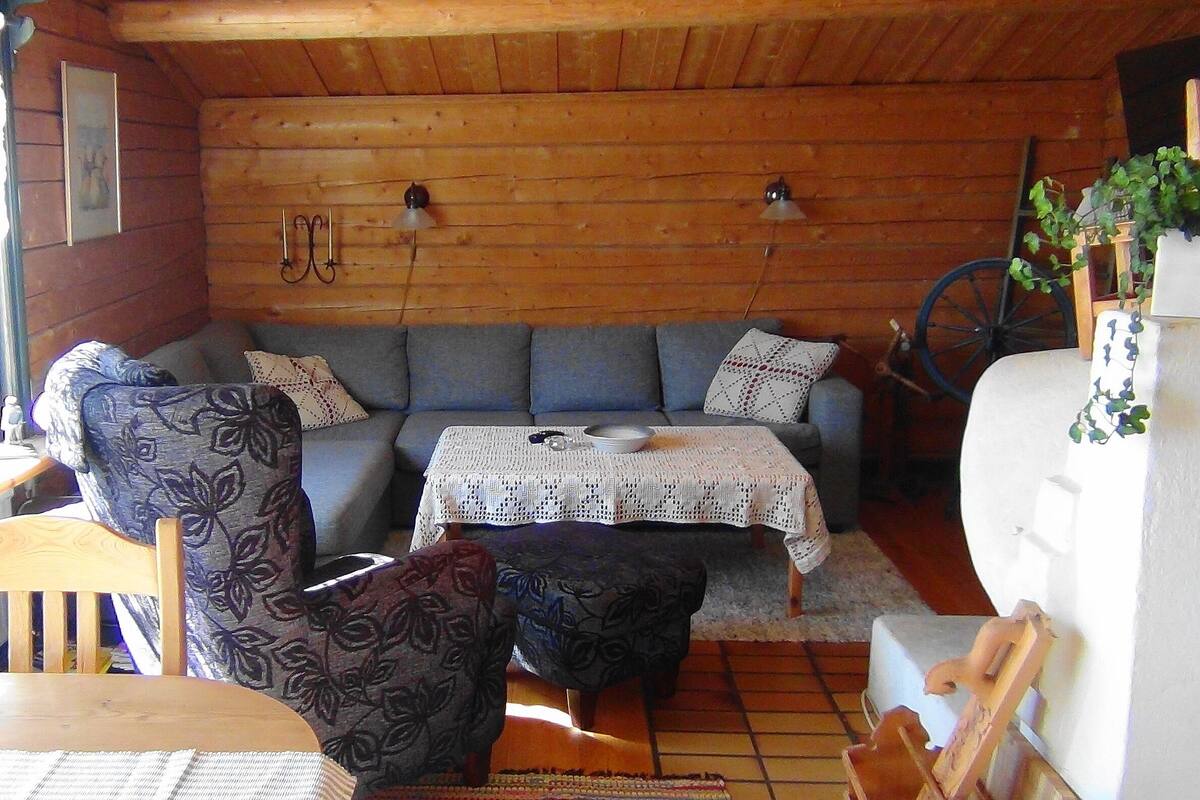 A cozy cabin interior with a wooden ceiling, a sofa, a patterned armchair, a small table, and a white tablecloth.