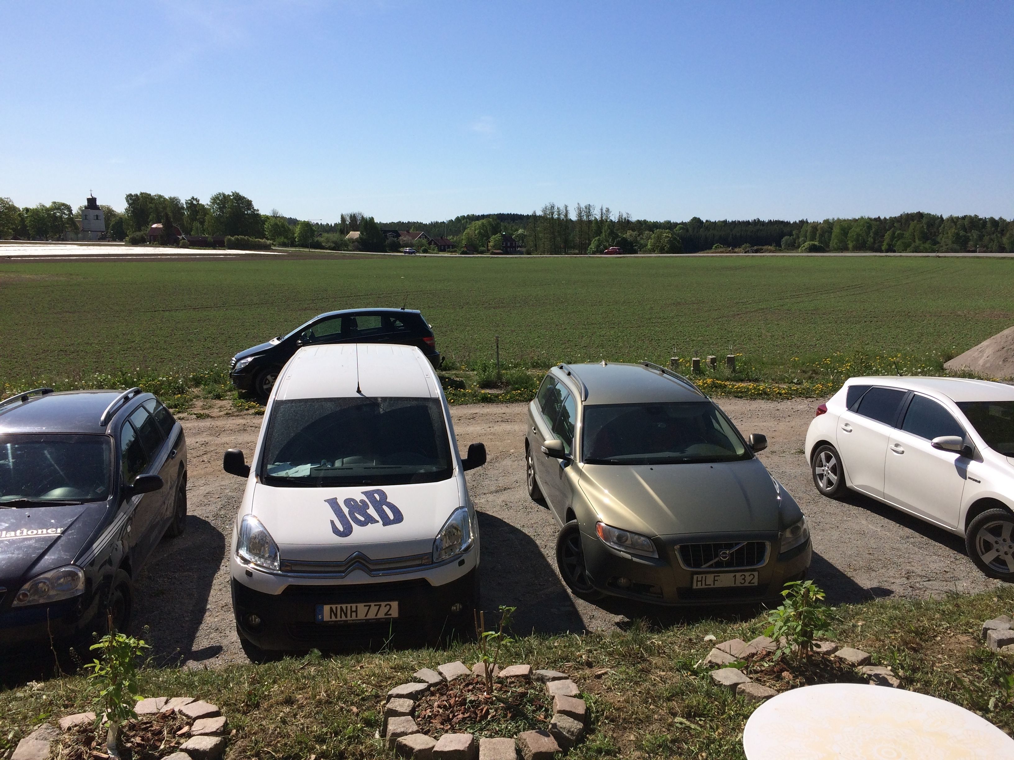 Cuatro coches estacionados en fila sobre un camino de tierra, con un campo cubierto de hierba y árboles al fondo.