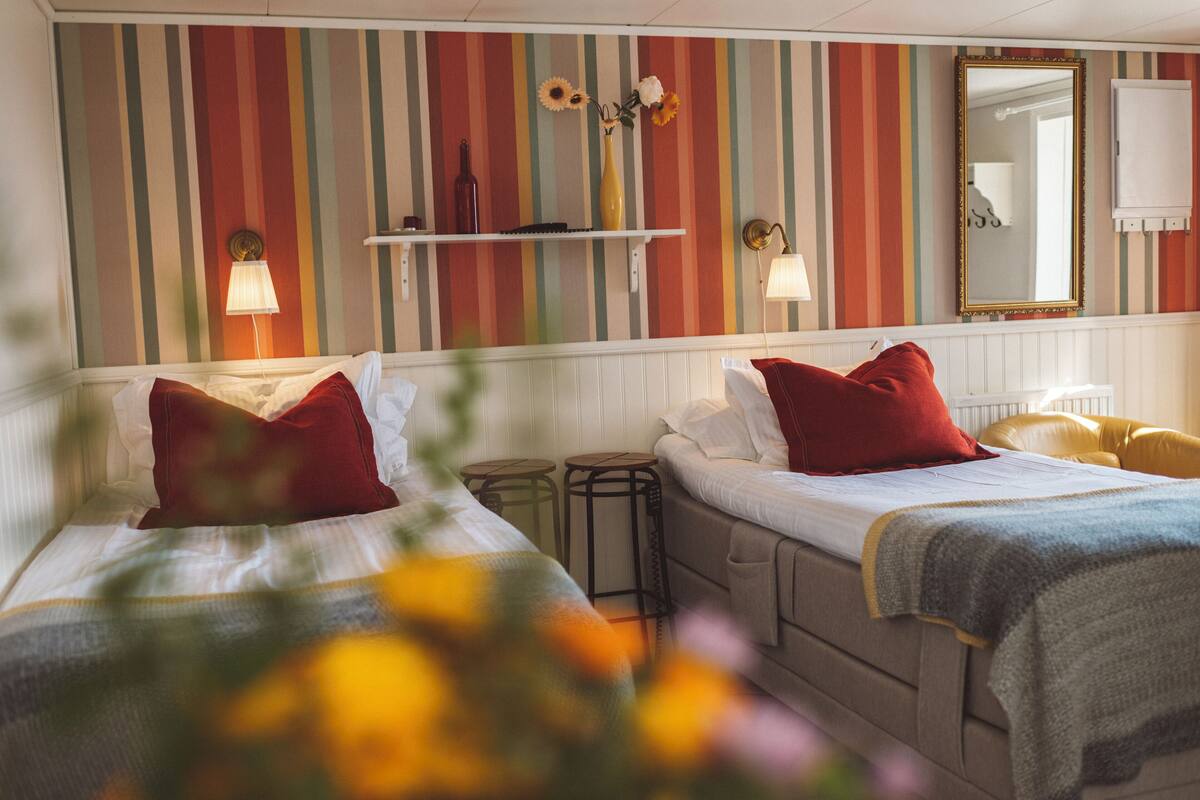 A hotel room with two beds, striped wallpaper, a mirror, and a shelf with decorative items.