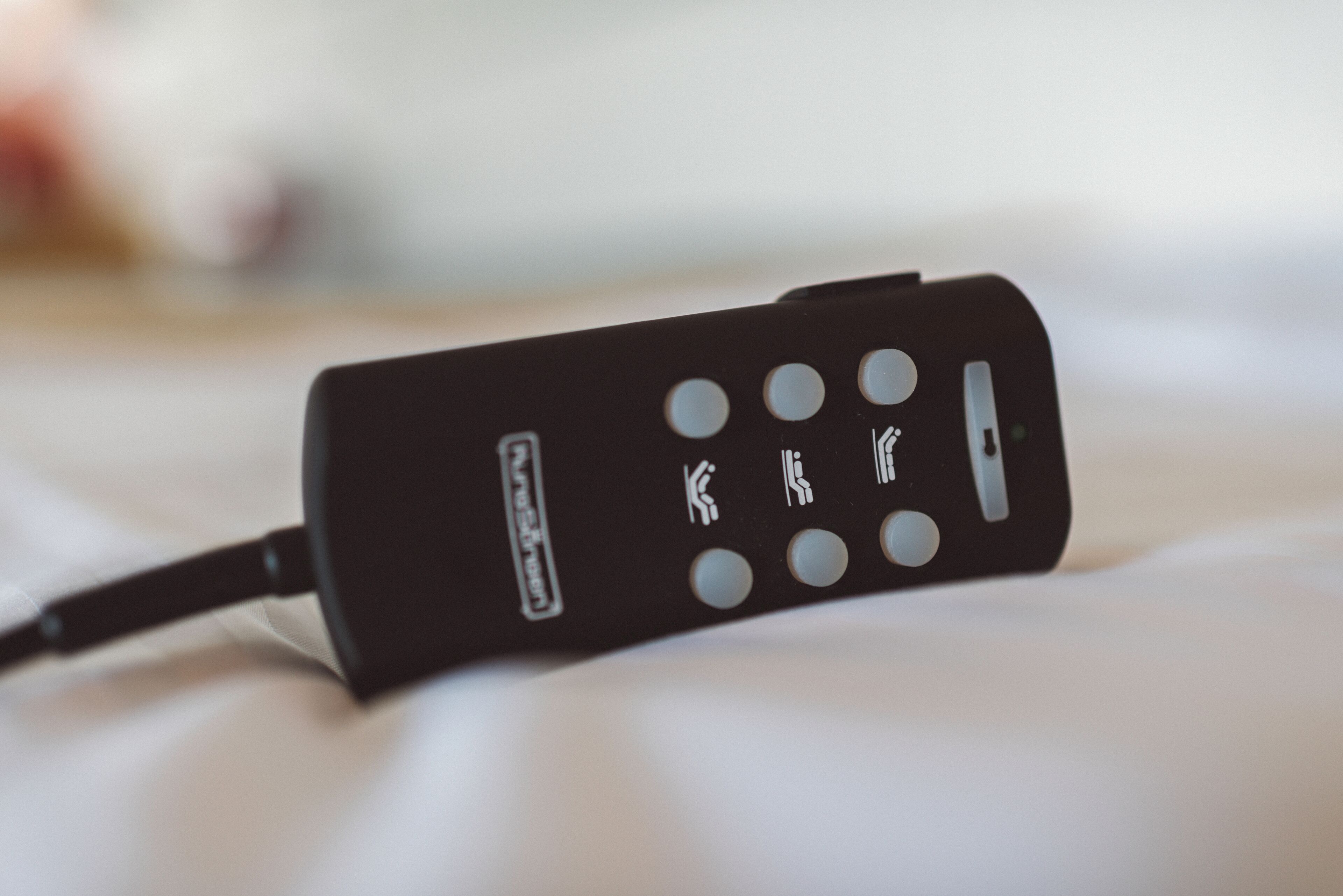 A black remote control with multiple buttons and a power cord, lying on a white surface.