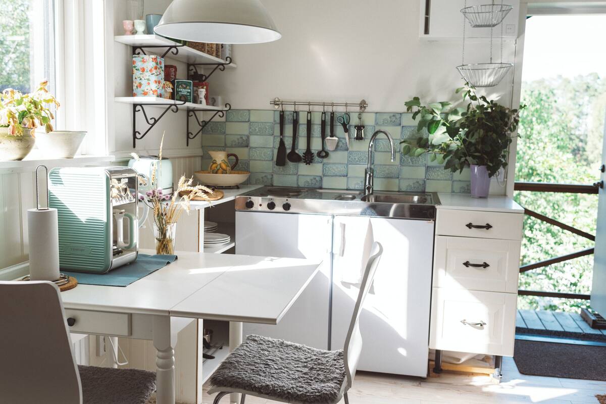 A compact kitchen with white cabinetry, a sink, and a stove. There are hanging utensils above the sink and a potted plant on the counter.