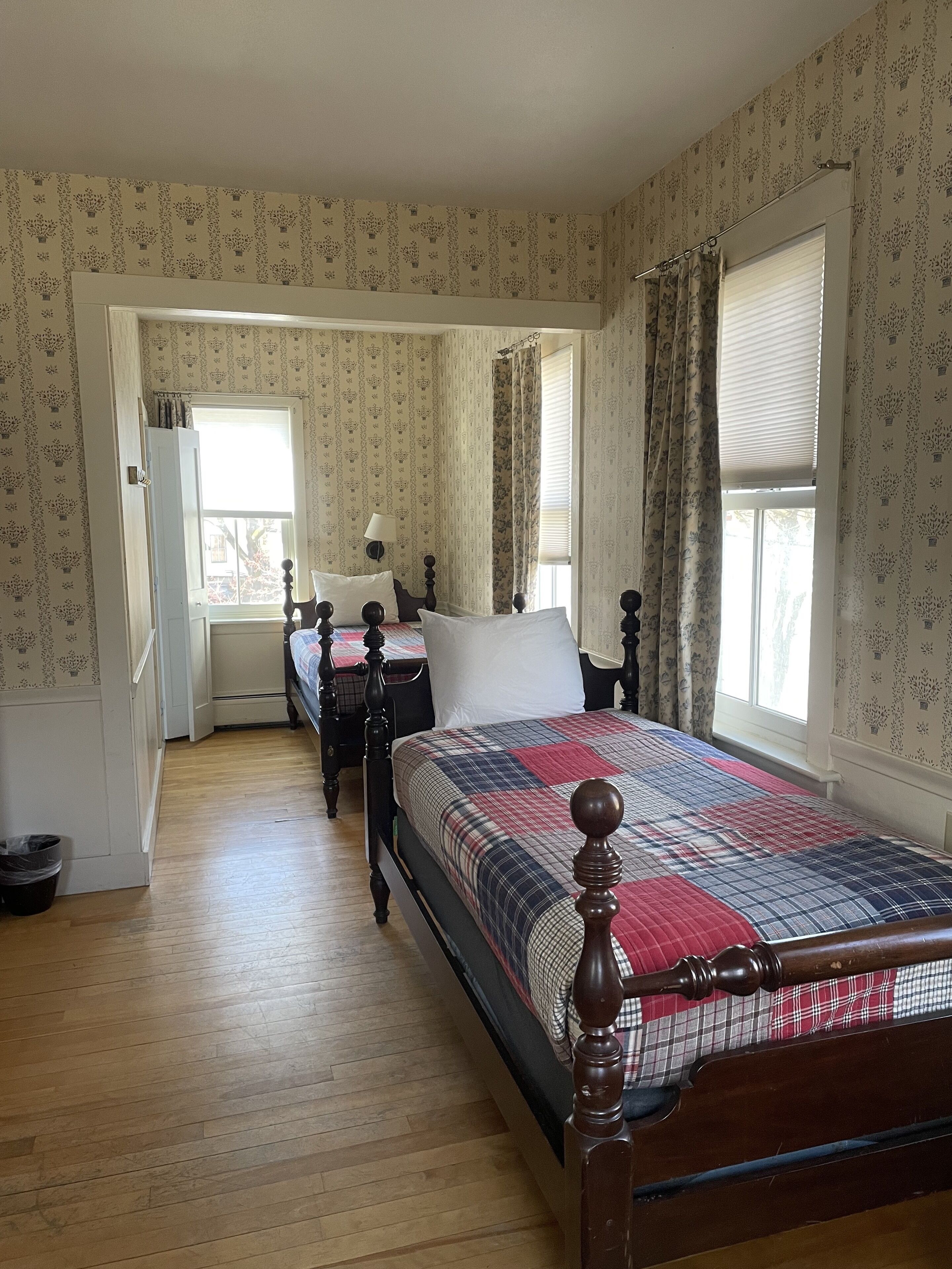 A bedroom with two beds, a wooden floor, patterned wallpaper, and a window with curtains.