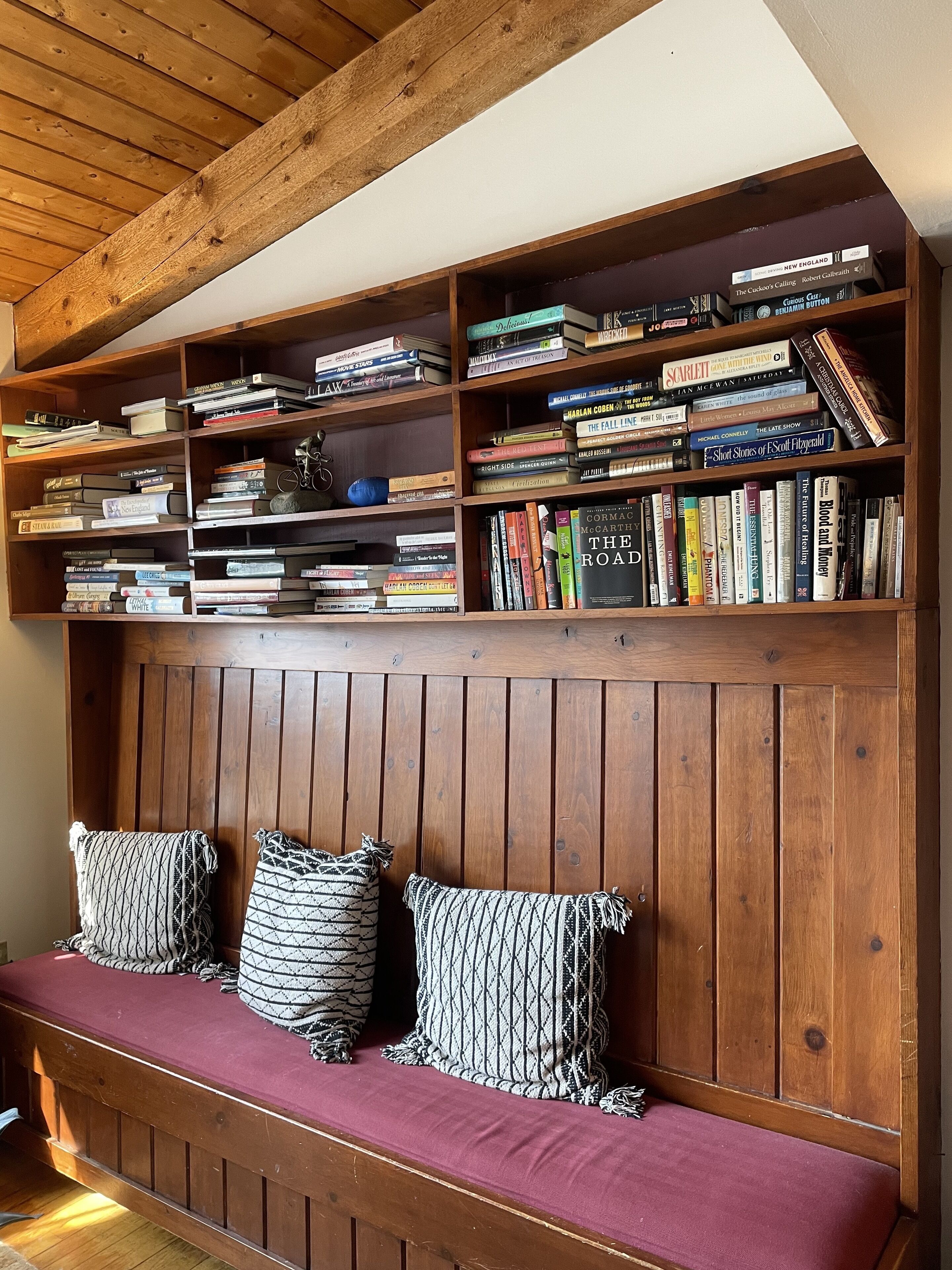 Una estantería de madera llena de libros, un banco de madera con cojines estampados y un techo de madera con vigas a la vista.