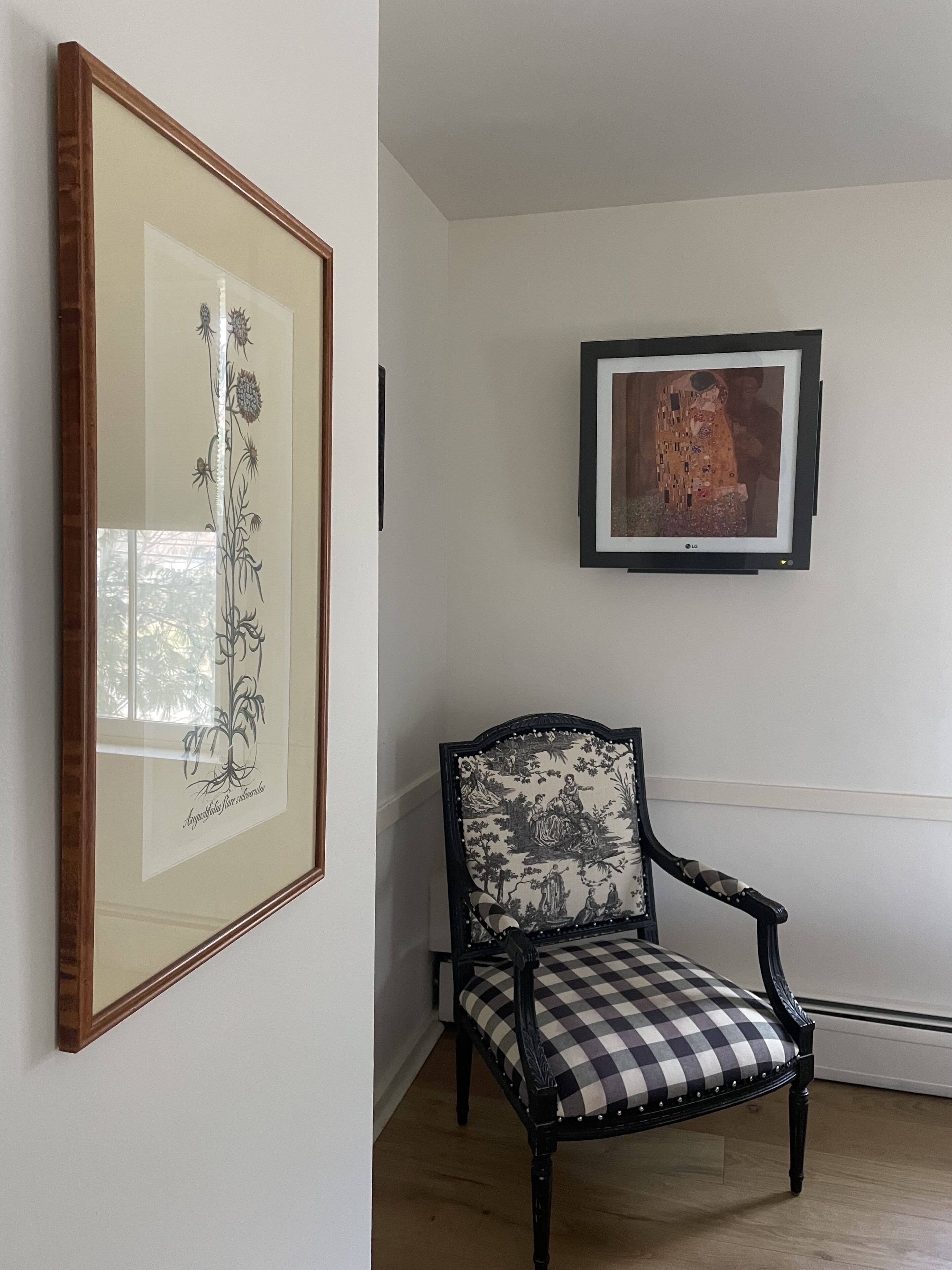 Une pièce décorée d’une estampe botanique encadrée, d’une œuvre d’art encadrée et d’un fauteuil à motifs à carreaux.