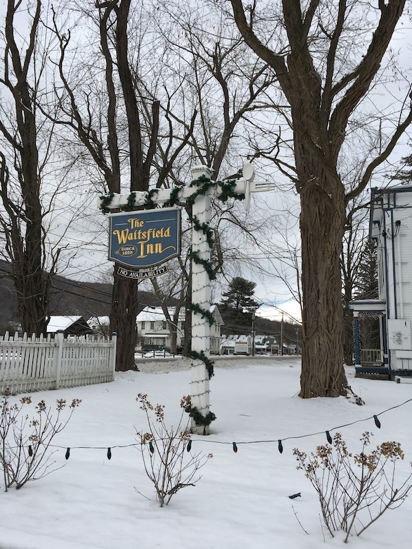 A sign for The Wainfield Inn with a white picket fence and bare trees in the background. - The Waitsfield Inn (Waitsfield)