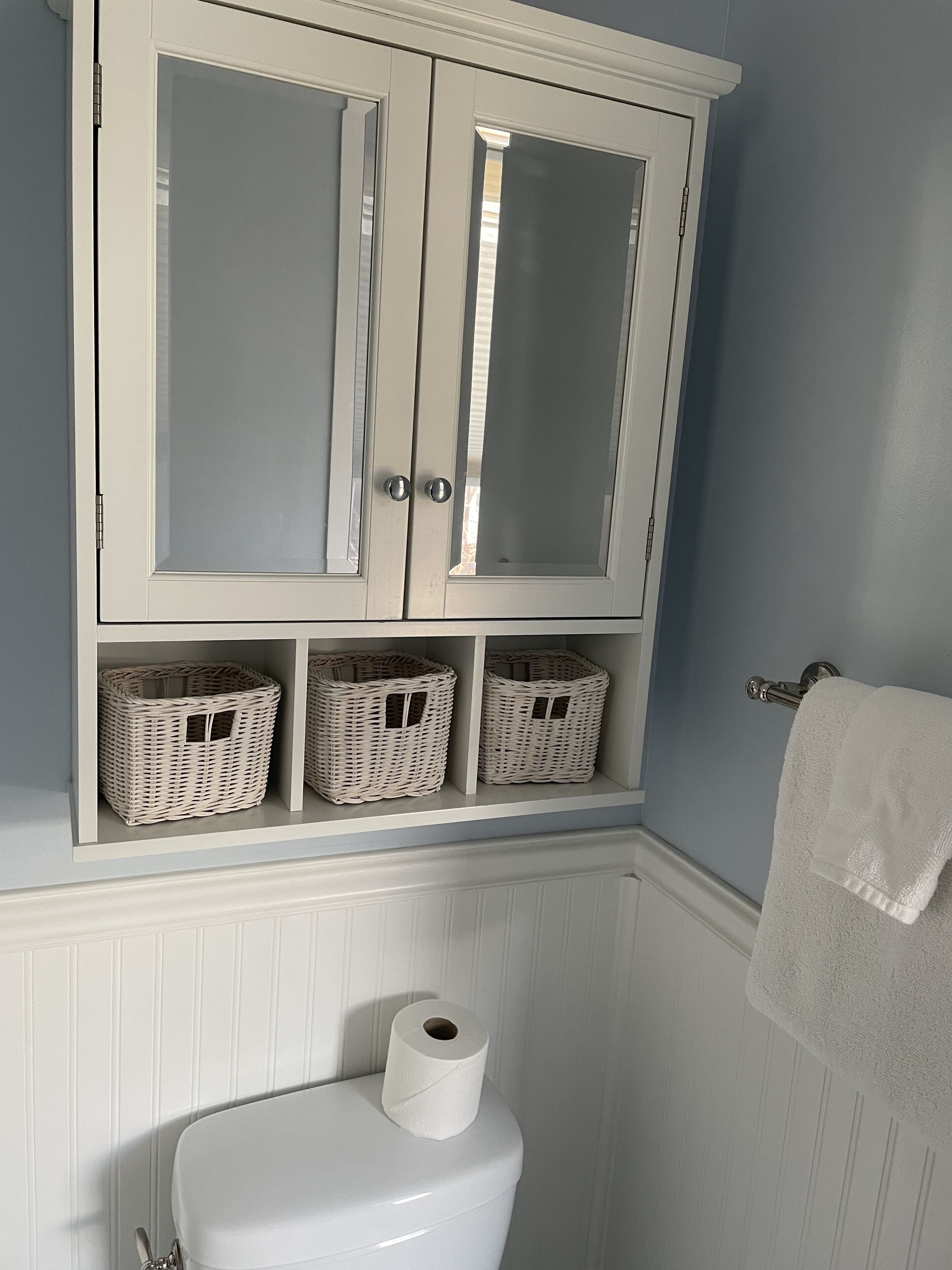 A bathroom with a white toilet, a roll of toilet paper, a white towel hanging on a towel rack, and three wicker baskets on a shelf above the toilet.