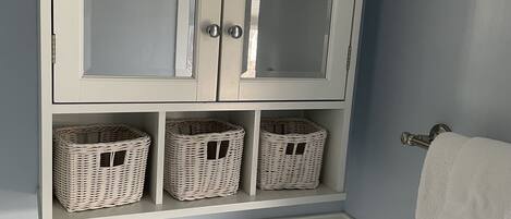 A bathroom with a white toilet, a roll of toilet paper, a white towel hanging on a towel rack, and three wicker baskets on a shelf above the toilet.