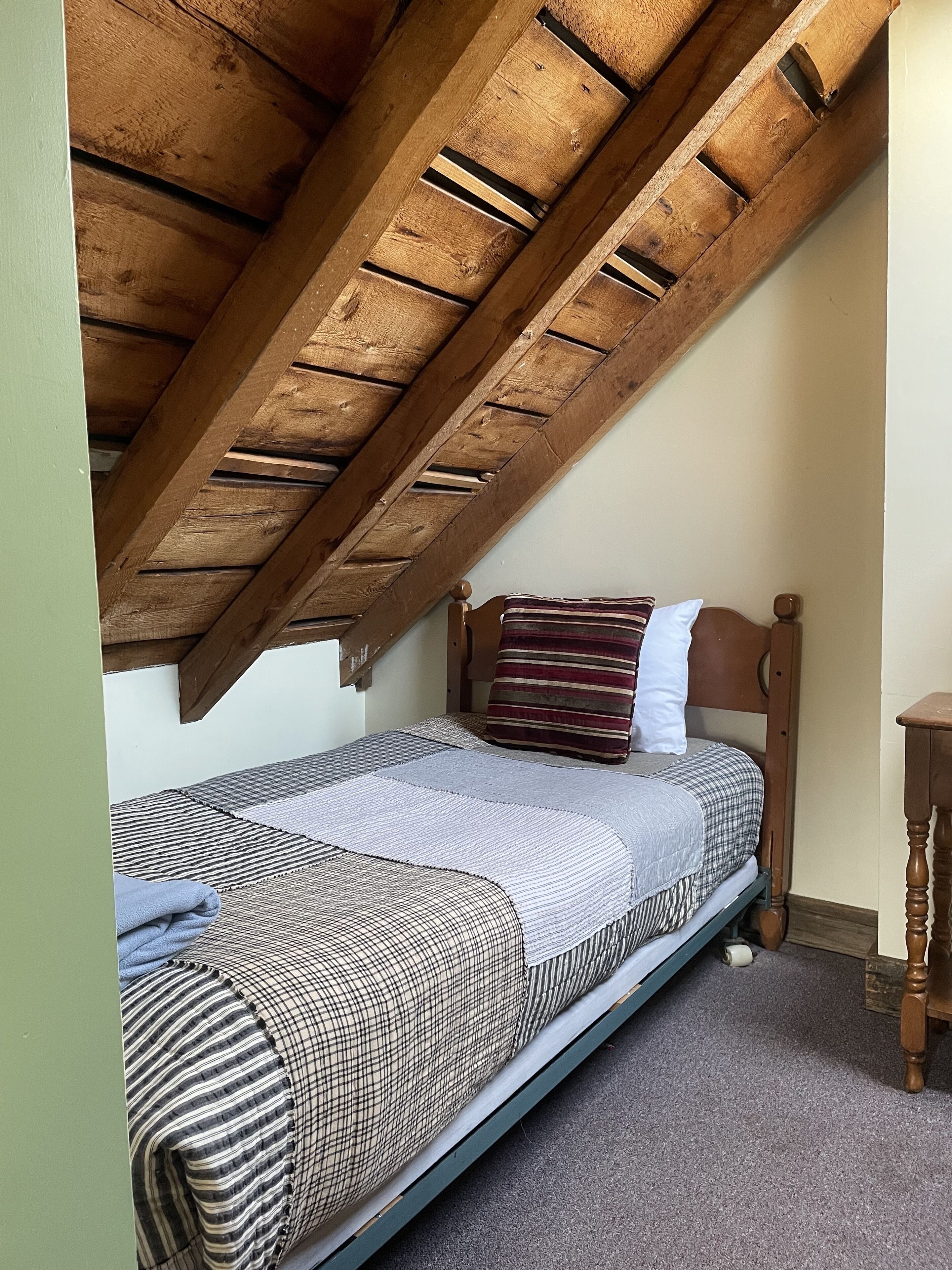 Una habitación pequeña con cama individual, techo de madera inclinado, cabecera de madera y una mesita de noche de madera.