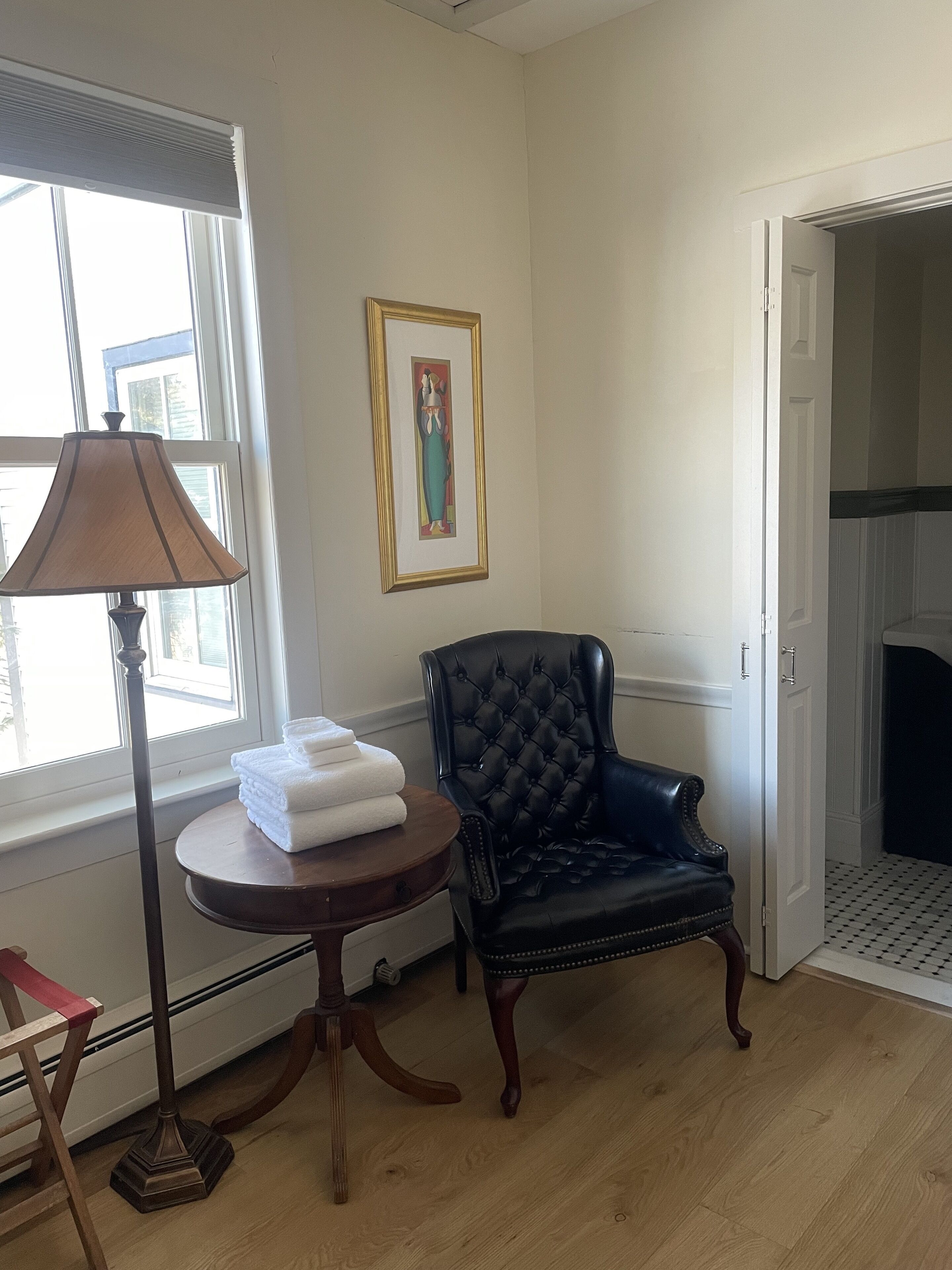 A room with a black leather armchair, a round wooden table with folded white towels, a floor lamp, and a framed artwork on the wall.