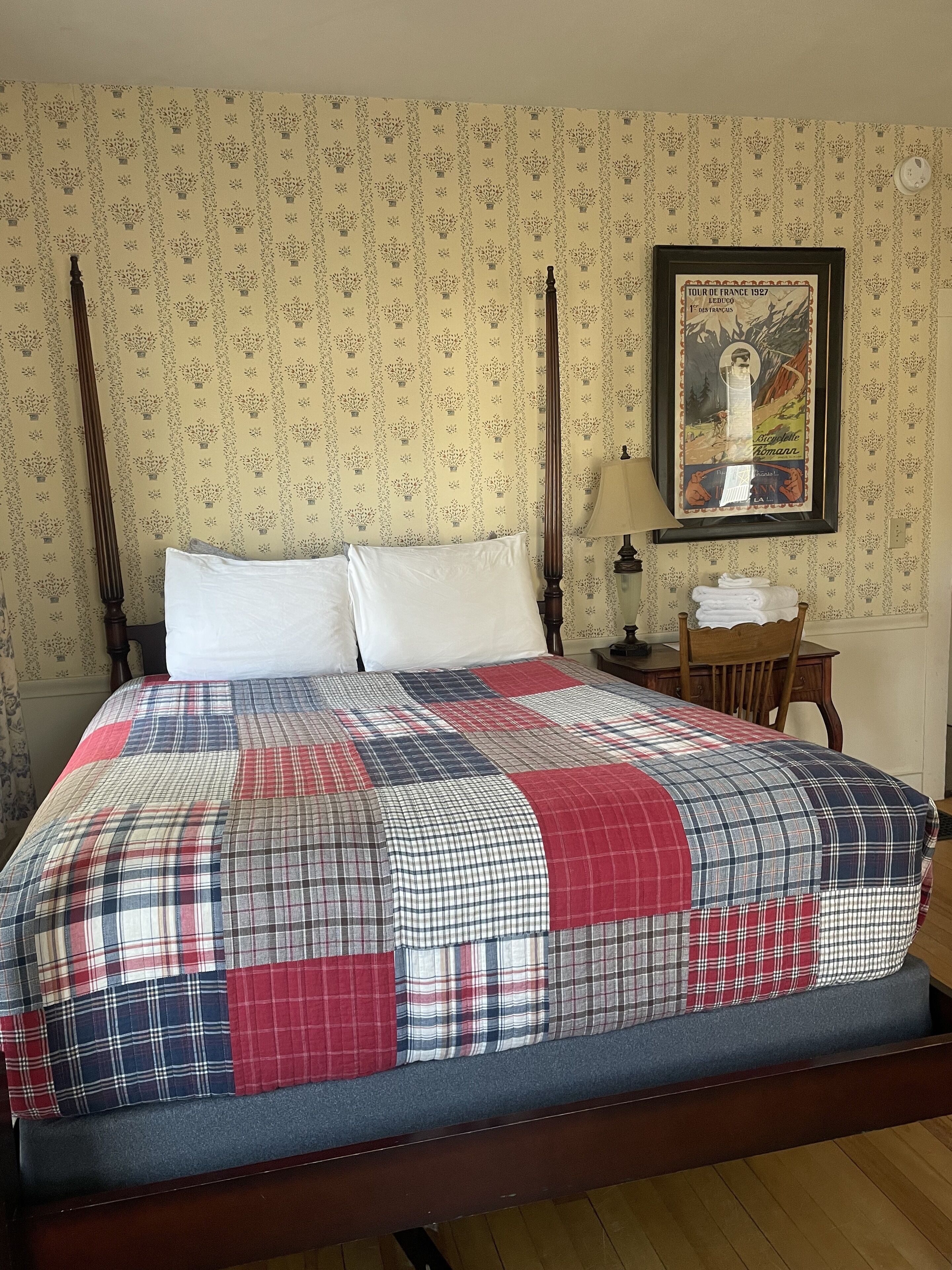 Una cama bien hecha con una colcha escocesa y dos almohadas, una mesita de noche de madera con una lámpara, un cartel enmarcado en la pared y una silla con toallas dobladas.