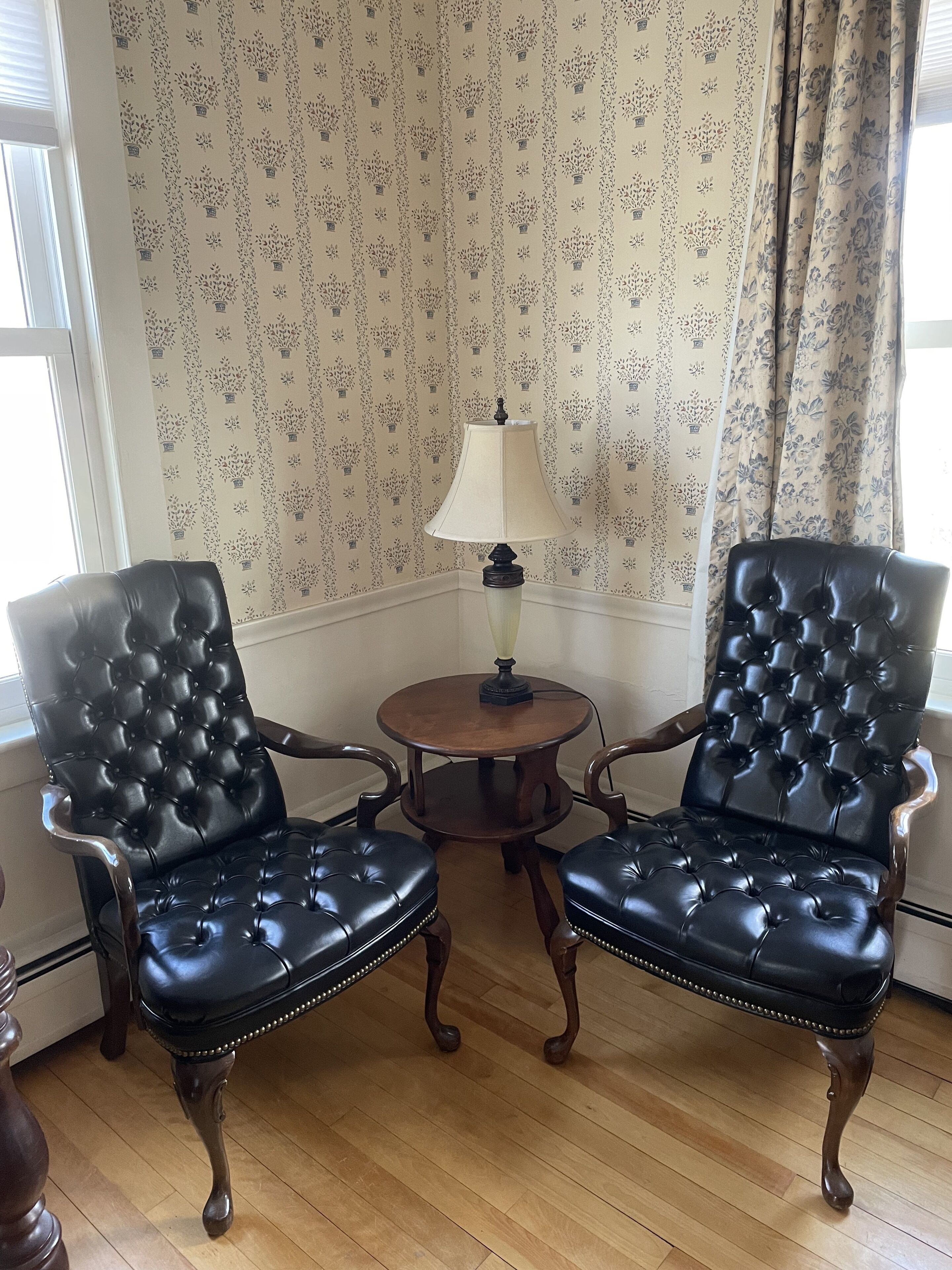 Dos sillones de cuero negro con una mesa en medio, una lámpara sobre la mesa y papel tapiz con motivos florales al fondo.