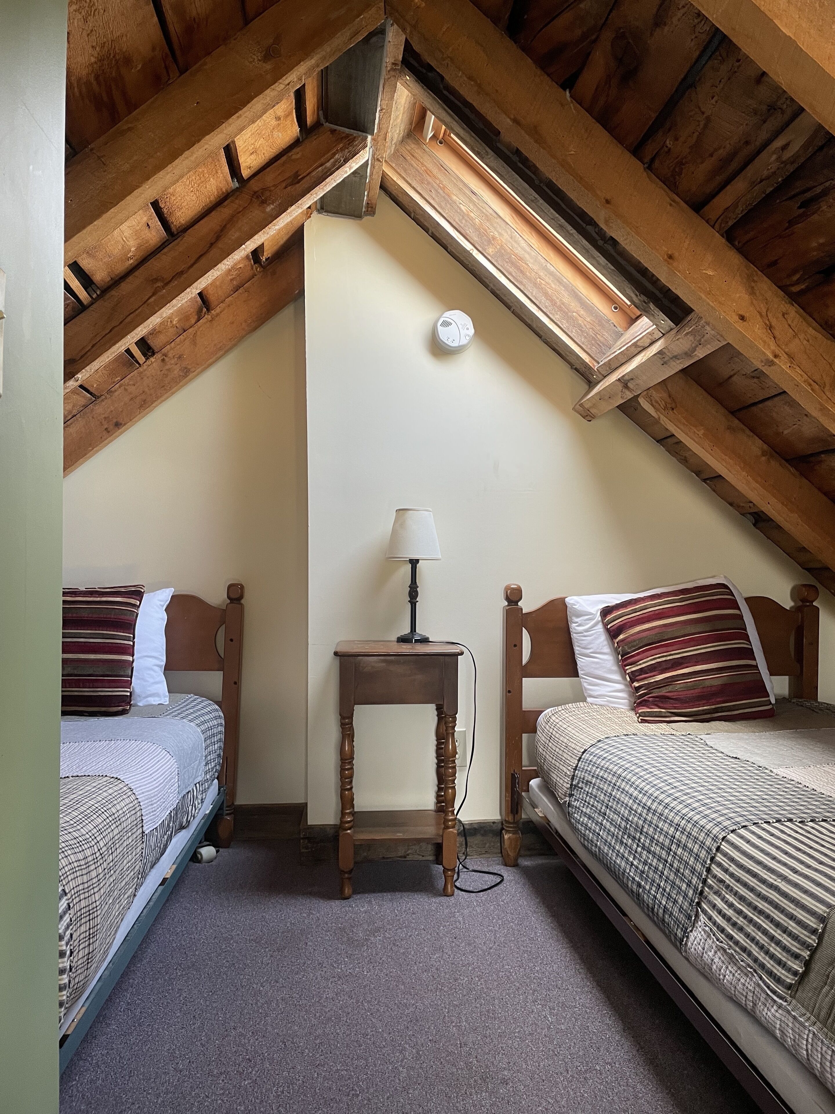 A small, cozy attic bedroom with two wooden beds, a nightstand, and a ceiling fan.