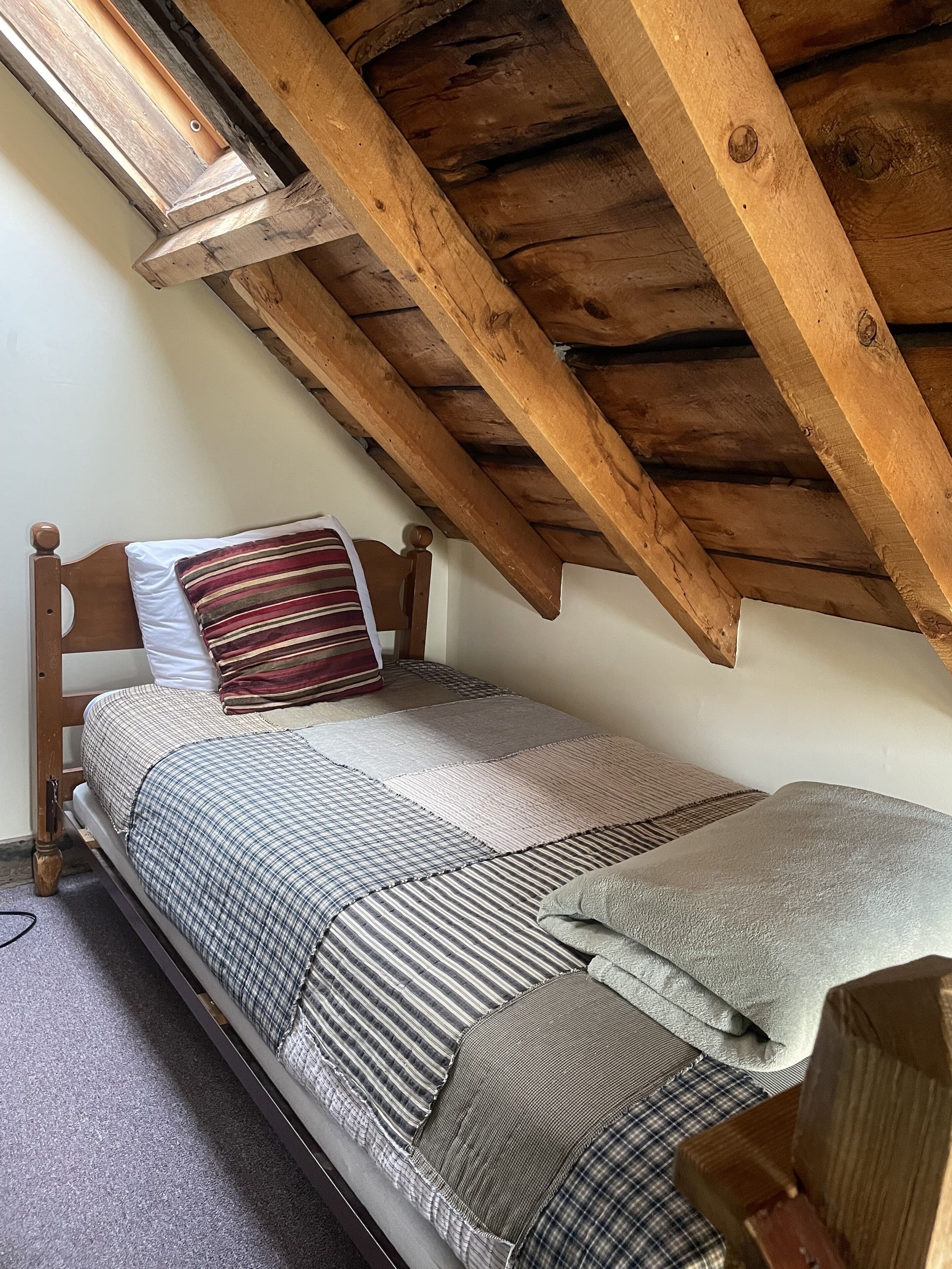 Ein kleiner, gemütlicher Raum mit schrägen Wänden und Holzbalken, mit einem Einzelbett, ausgestattet mit vielen Kissen und einer gestreiften Decke.
