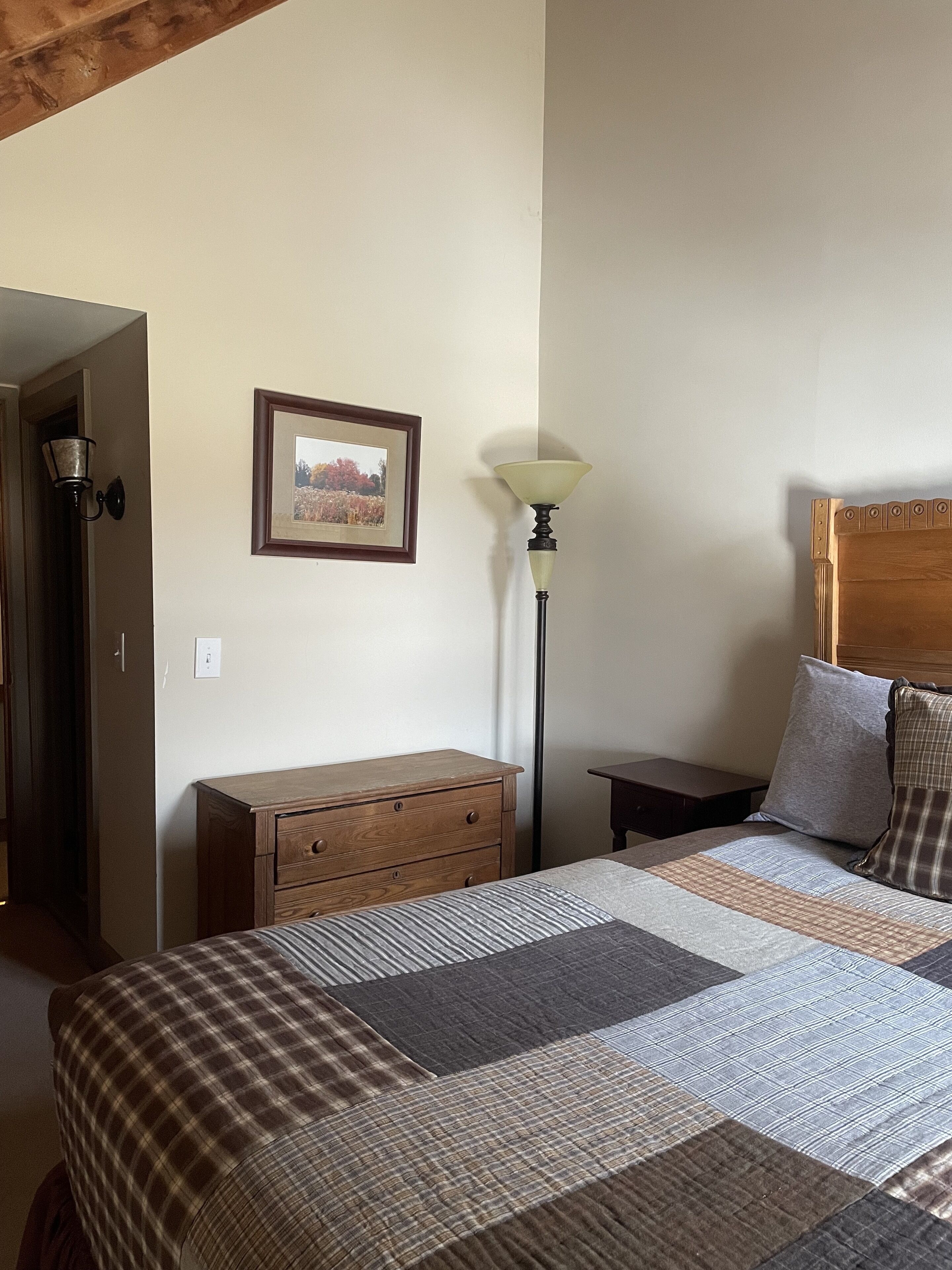 Une chambre à coucher avec un lit, une commode en bois, une table de chevet, une lampe et un tableau encadré accroché au mur.