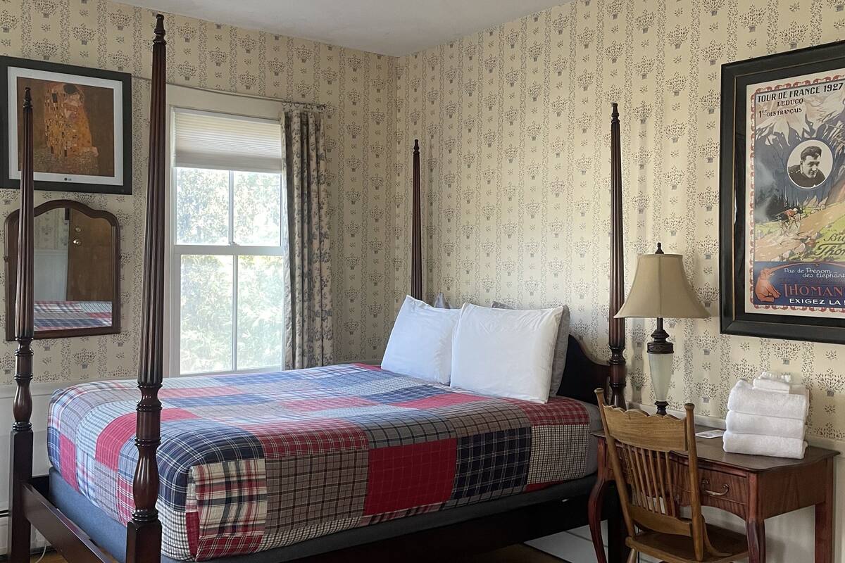 Una cama con dosel con una colcha escocesa, una silla de madera, un escritorio con una lámpara y un cuadro en la pared.