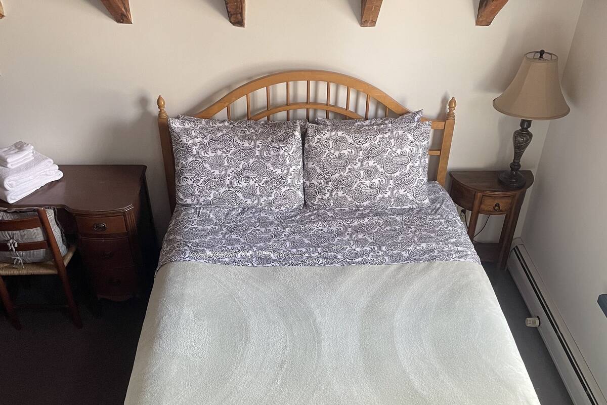 Una cama bien tendida con ropa de cama estampada, un cabecero de madera, una mesita de noche con una lámpara y una silla con cojín.