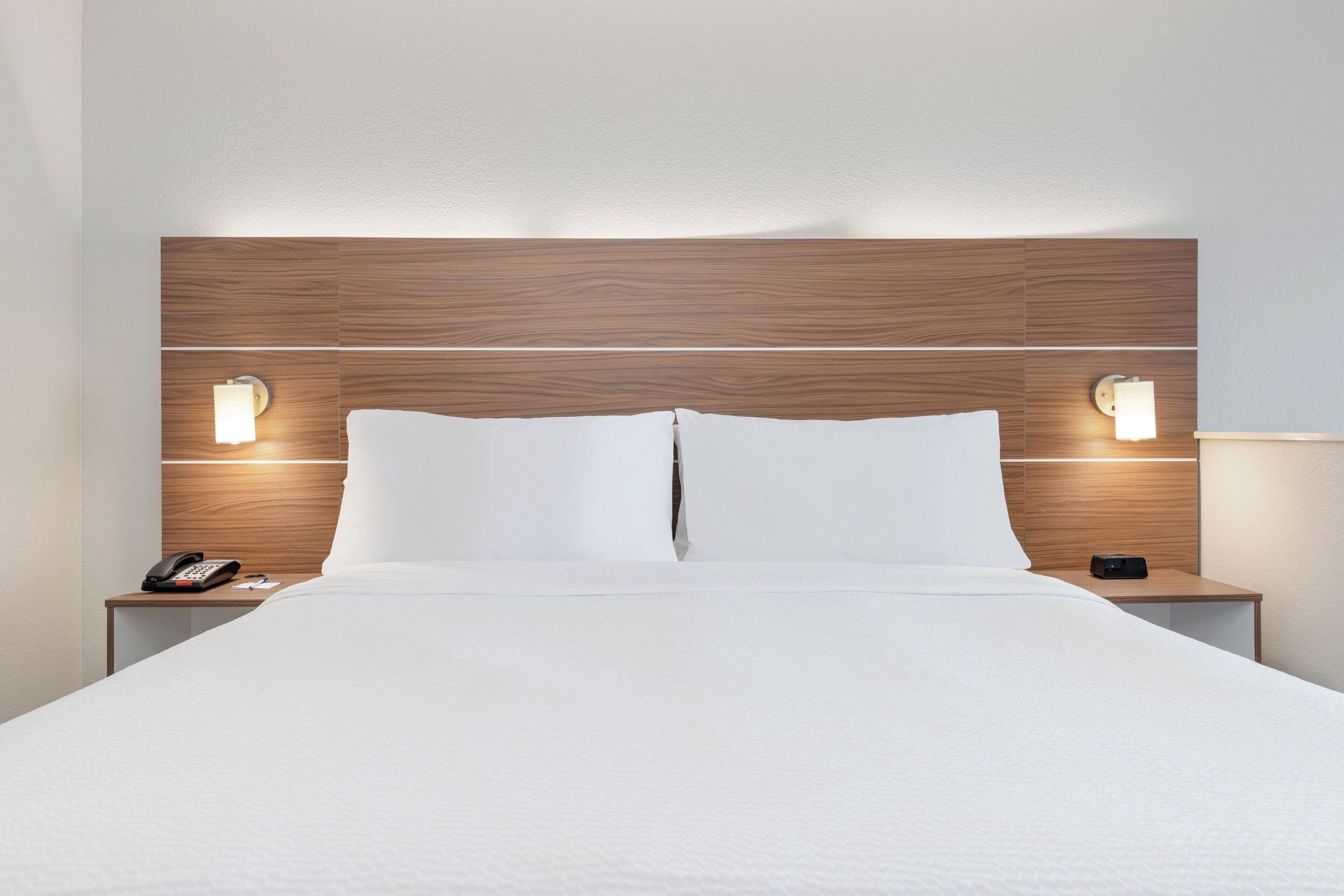 A hotel room with a wooden headboard, two bedside lamps, a white bedspread, and a nightstand with a phone and a remote control.