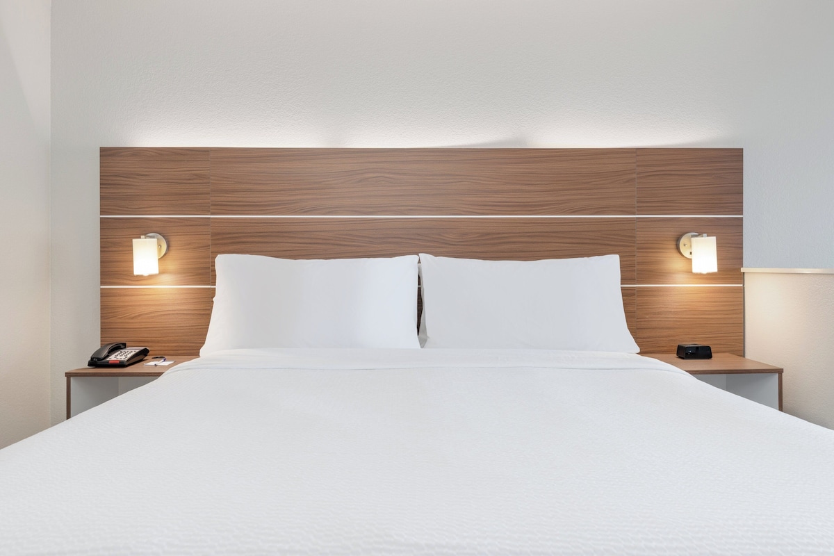 Une chambre d’hôtel avec une tête de lit en bois, deux lampes de chevet, une couvre-lit blanche et une table de chevet avec un téléphone et une télécommande.