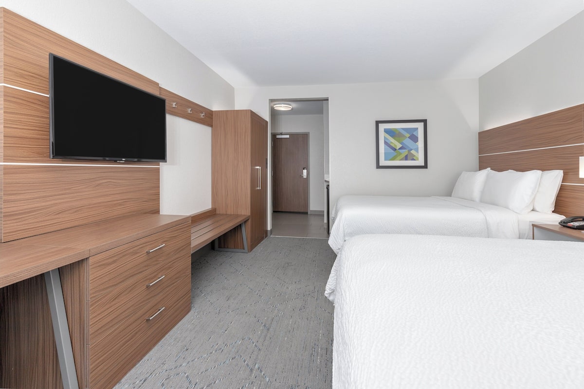 A hotel room with a TV mounted on the wall, a desk with drawers, a bed with white bedding, and a door leading to another room.